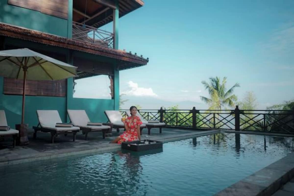 A woman sitting by a pool with lounge chairs and umbrellas, a building with a balcony, and palm trees in the background.