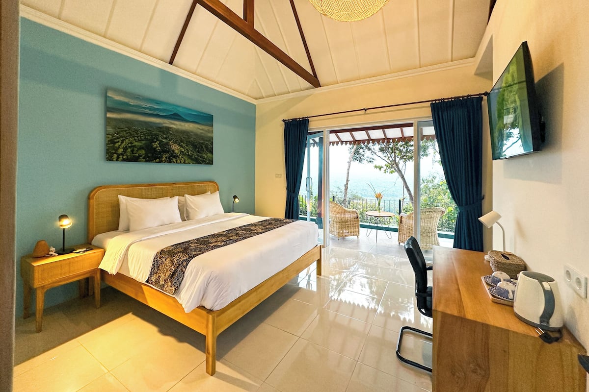 A bedroom with a bed, a nightstand, a desk with a lamp, a TV, and a view of the outdoors.