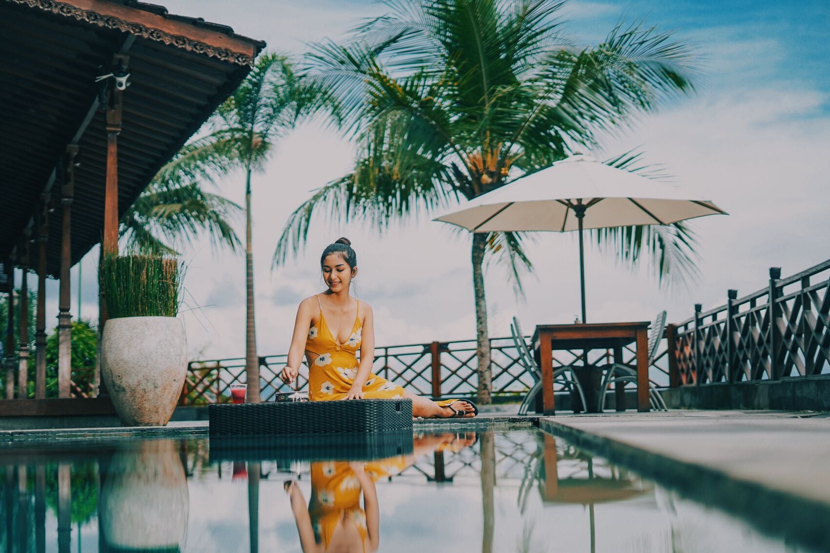 Una mujer sentada junto a una piscina, con una palmera y un paraguas grande al fondo.