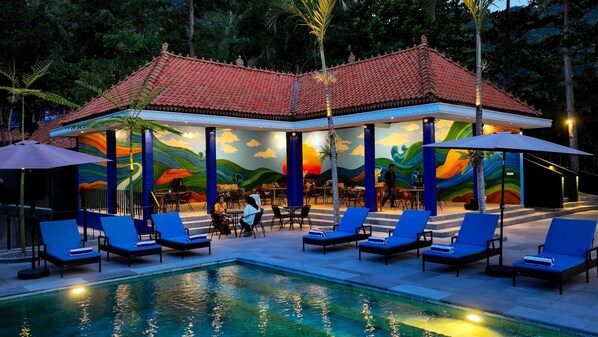 Un área de descanso junto a la piscina con sillones azules, una piscina y un edificio con tejado y murales.