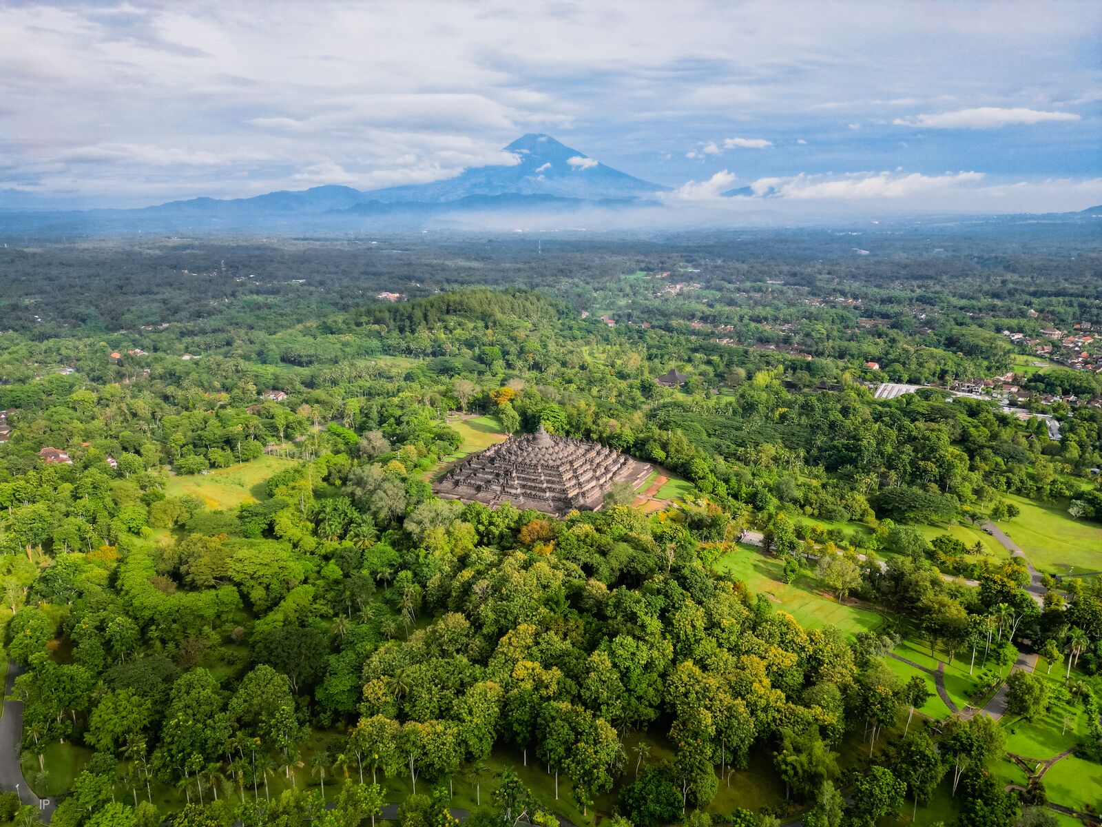 Une vue aérienne d’un paysage verdoyant et luxuriant, avec une structure en forme de pyramide entourée d’une végétation dense et une chaîne de montagnes à l’horizon.