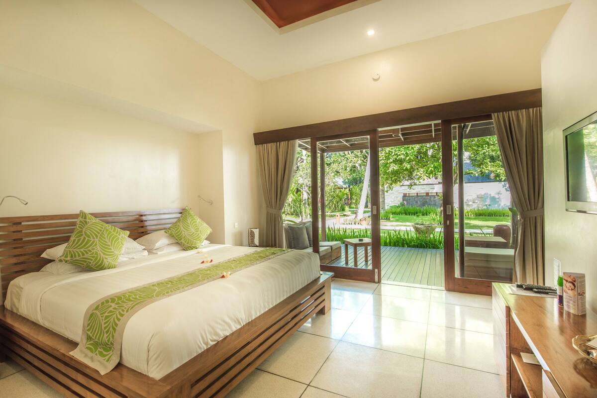 A spacious bedroom with a large bed, a wooden headboard, and a view of a lush garden through a sliding glass door.