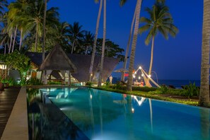 A swimming pool with a thatched-roof structure, palm trees, and a view of the ocean at dusk. - The Chandi Boutique Resort (Senggigi)