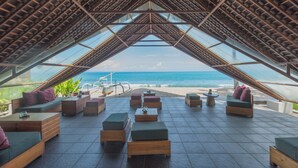 Un salón junto a la playa con vista al océano, con mobiliario de madera y una estructura de techo de cristal.
