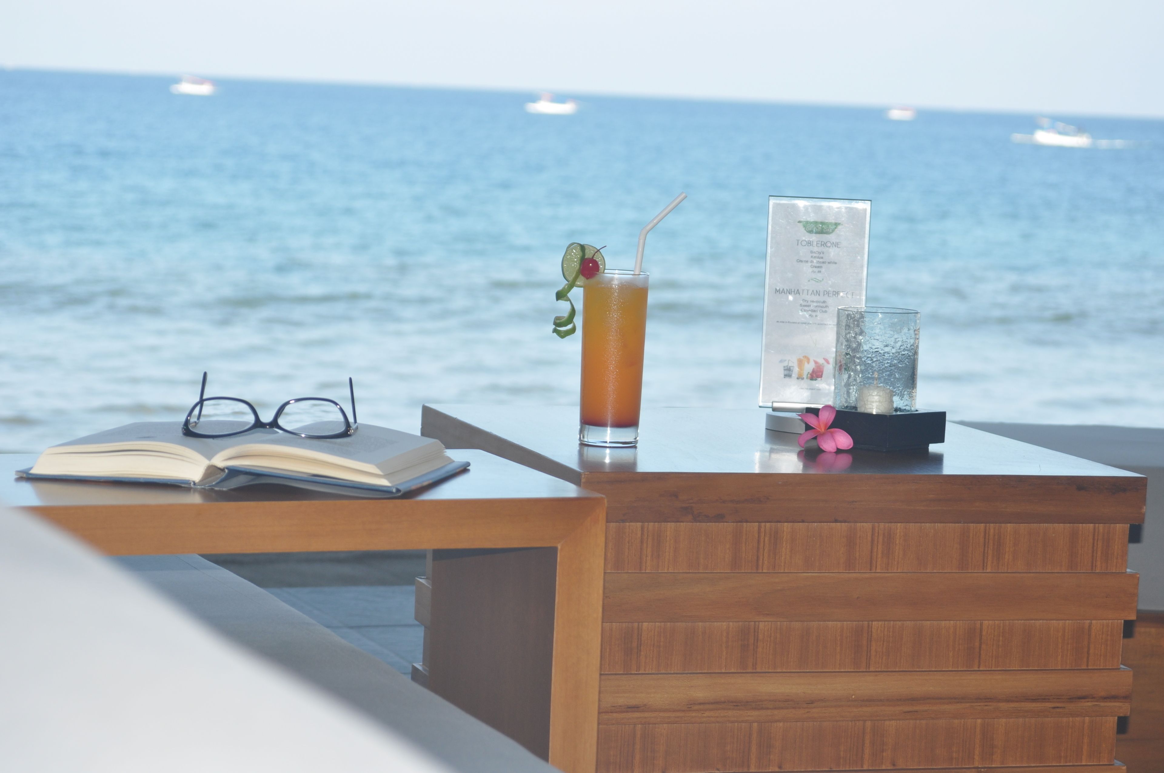 A wooden desk with an open book, a drink with a straw and cherry, and a menu on a seaside view.