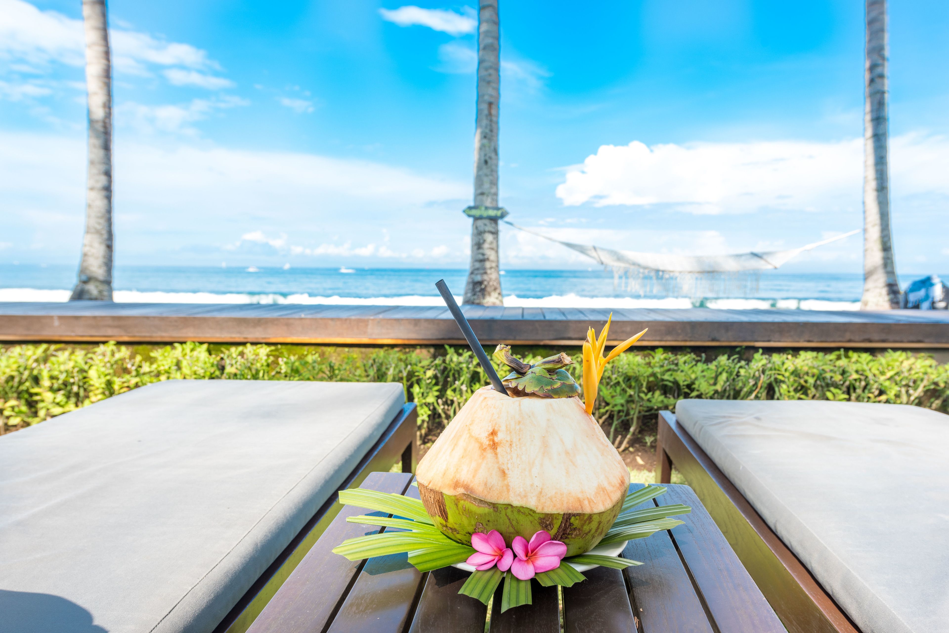 Une table en bois, sur laquelle se trouvent une boisson au lait de coco et une fleur rose, avec vue sur une plage bordée de palmiers et agrémentée d’un hamac.
