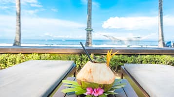 Une table en bois, sur laquelle se trouvent une boisson au lait de coco et une fleur rose, avec vue sur une plage bordée de palmiers et agrémentée d’un hamac.