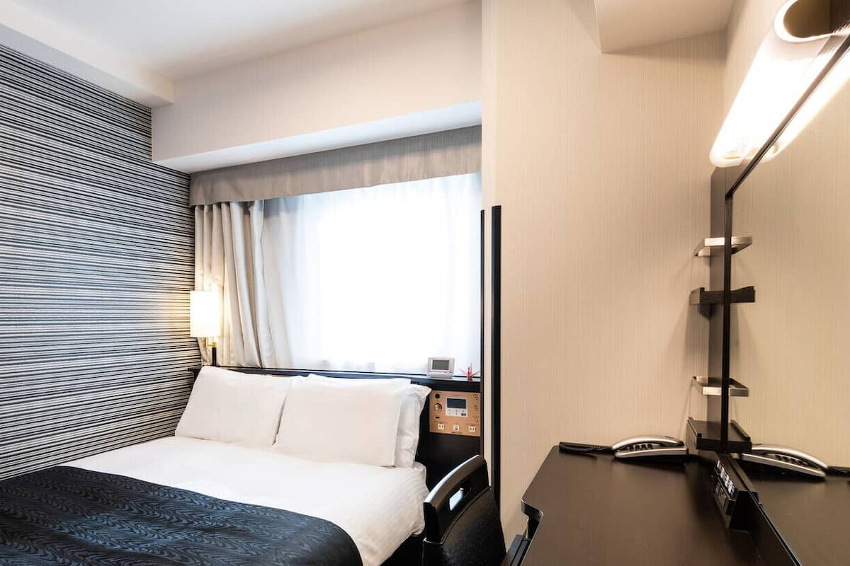 A hotel room with a bed, bedside table, lamp, and window with curtains.