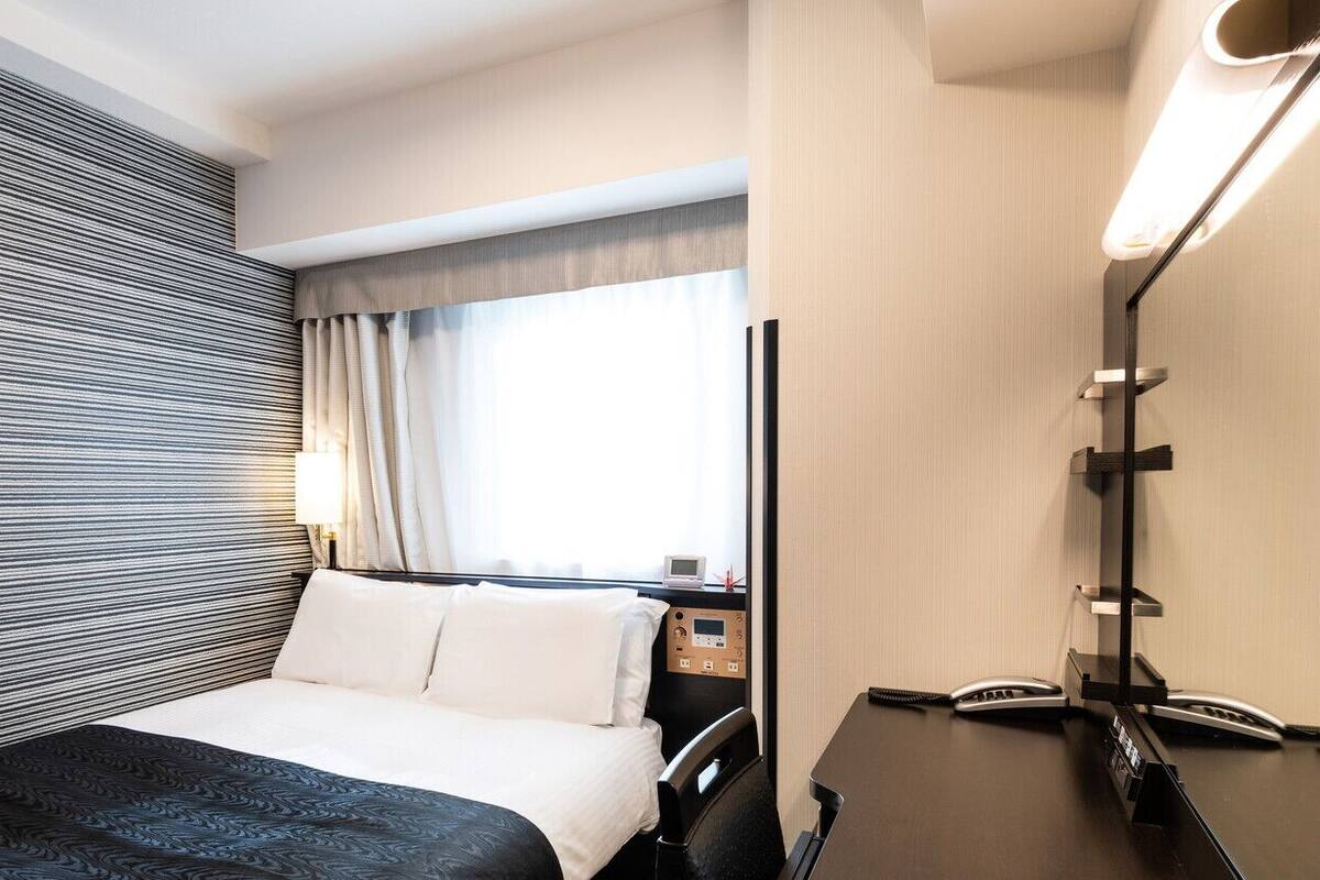 Habitación de hotel con cama, mesita de noche, lámpara y ventana con cortinas.