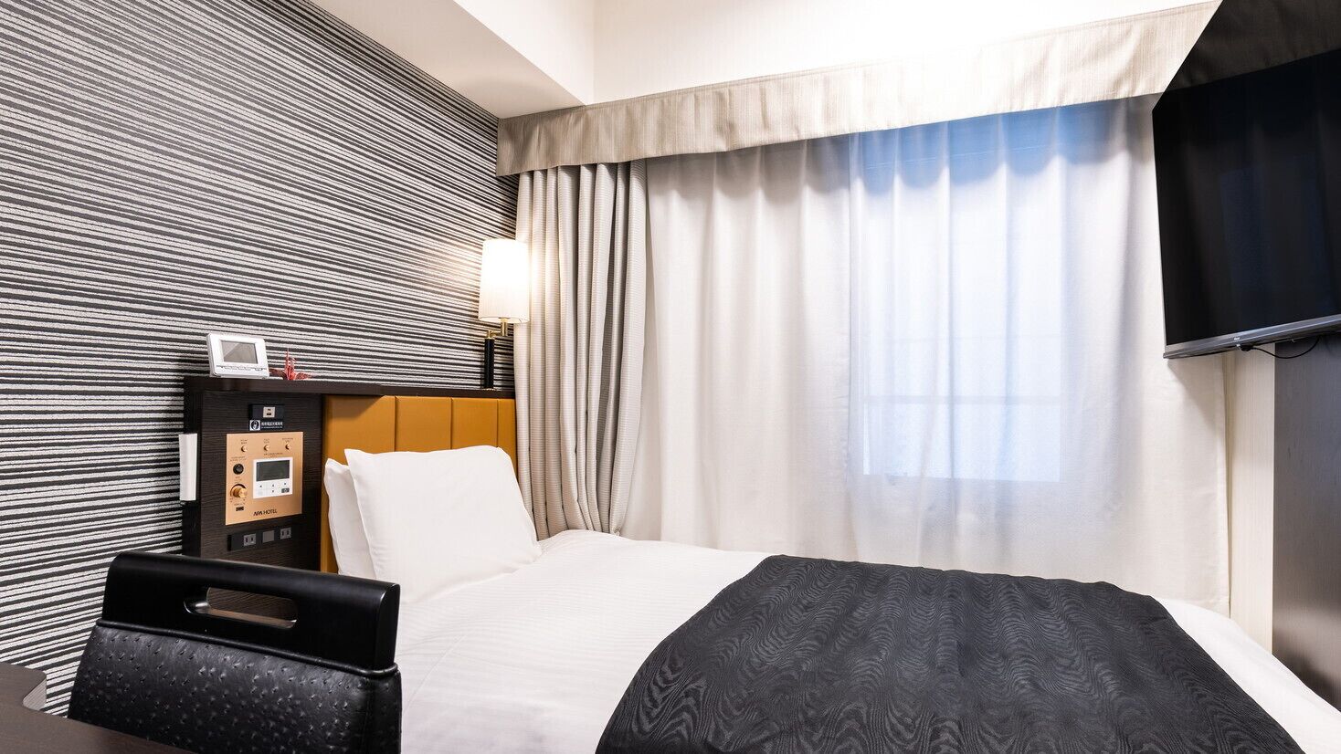 Una habitación de hotel con cama, panel de control, televisor y ventana con cortinas.
