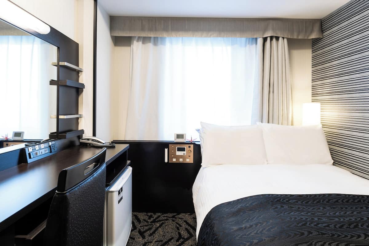 A hotel room with a bed, a desk with a phone, a mini-fridge, a control panel, and a window with curtains.