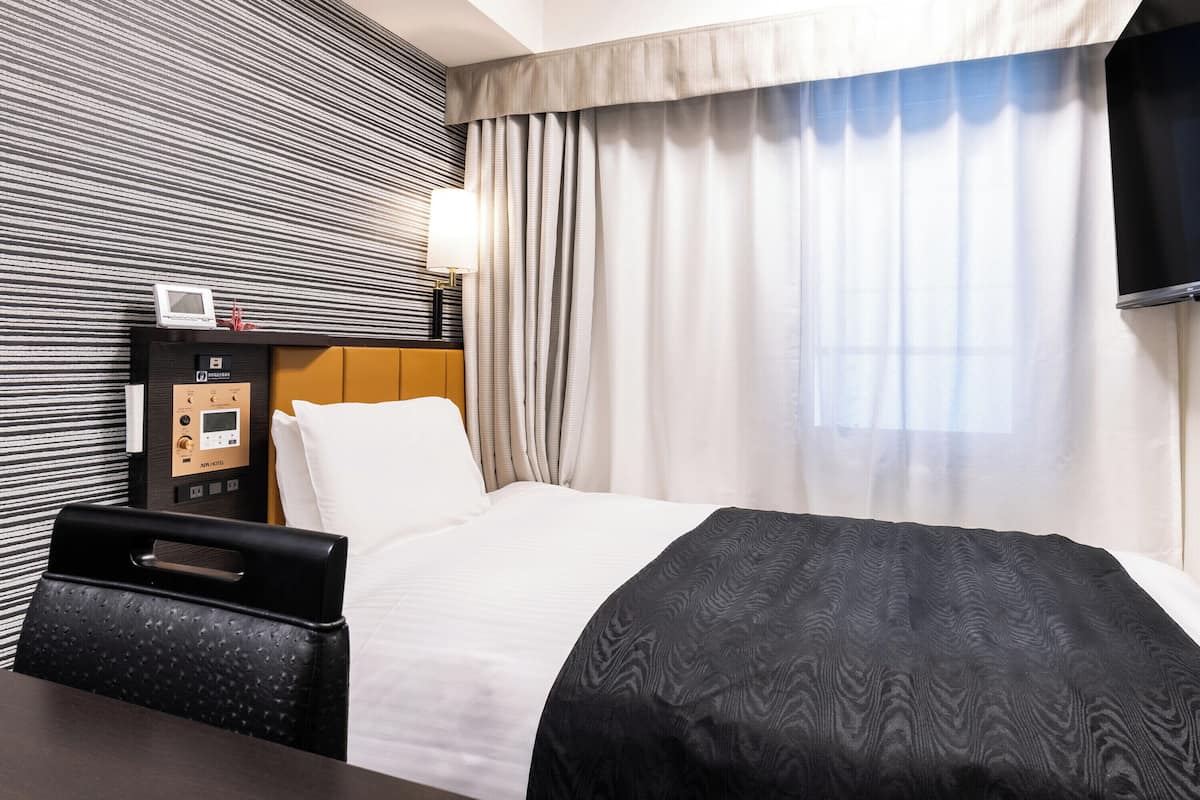 A hotel room with a bed, a desk, a lamp, a TV, and a control panel.