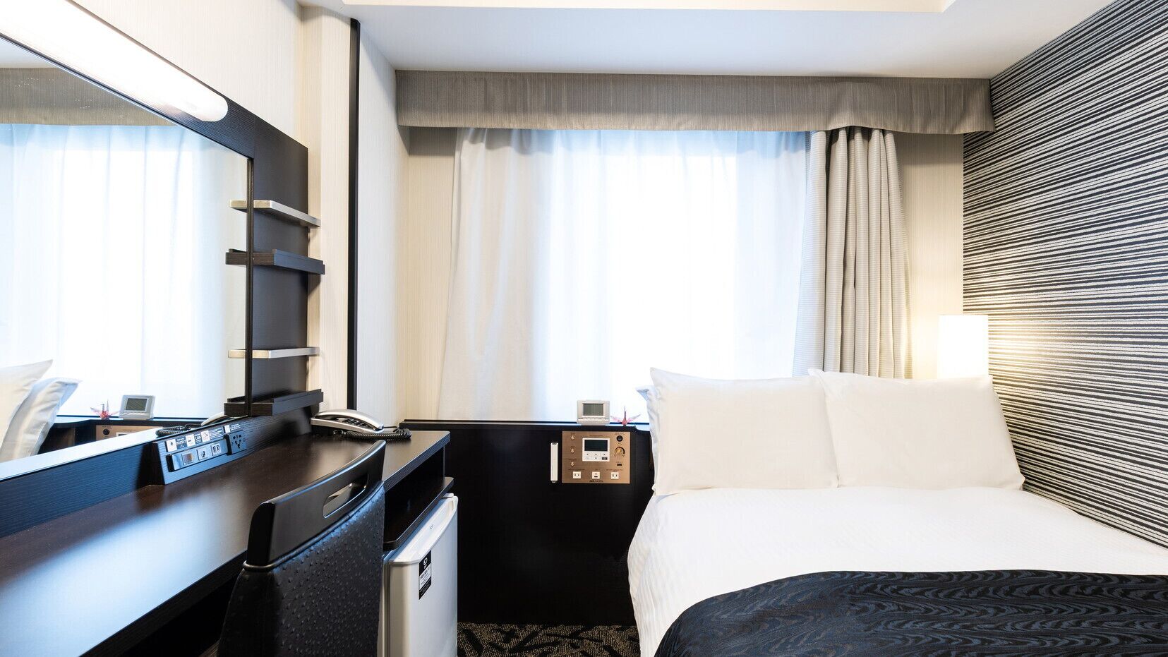 Habitación de hotel con cama, escritorio, espejo, mesita de noche, lámpara y ventana con cortinas.