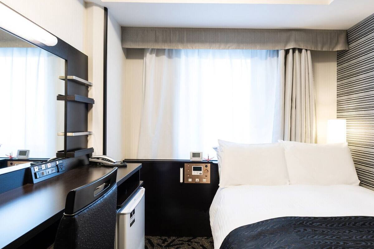 A hotel room with a bed, a desk, a mirror, a nightstand, a lamp, and a window with curtains.