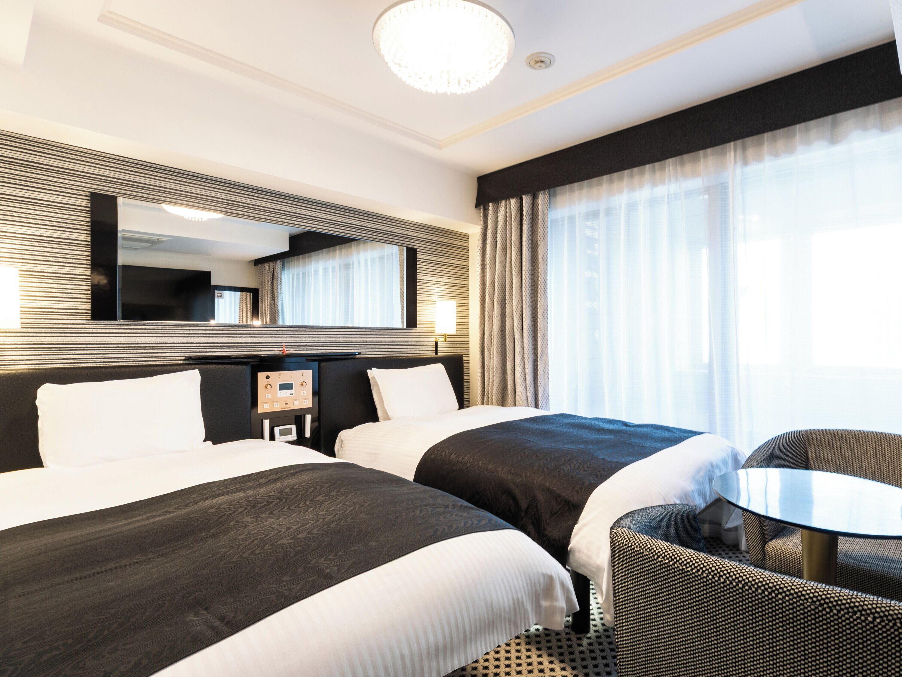 A hotel room with two beds, a small table, a chair, a large mirror, and a window with curtains.