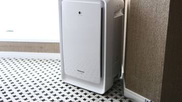 A white portable air purifier in a corner of a room with a patterned floor and a window in the background.