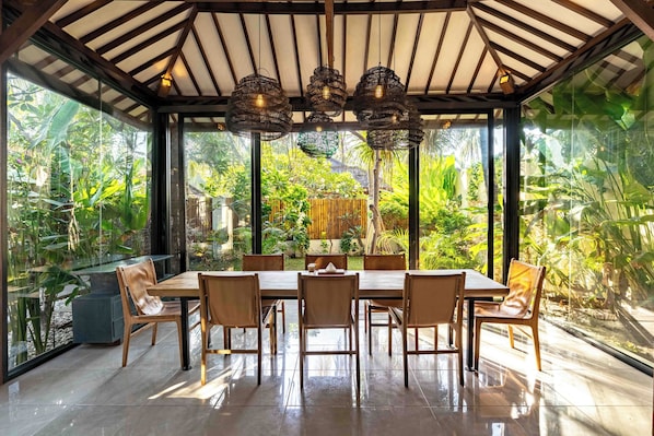 A dining area with a glass enclosure, a wooden table, and chairs, surrounded by lush greenery. - Kelapa Villas (Gili Trawangan)