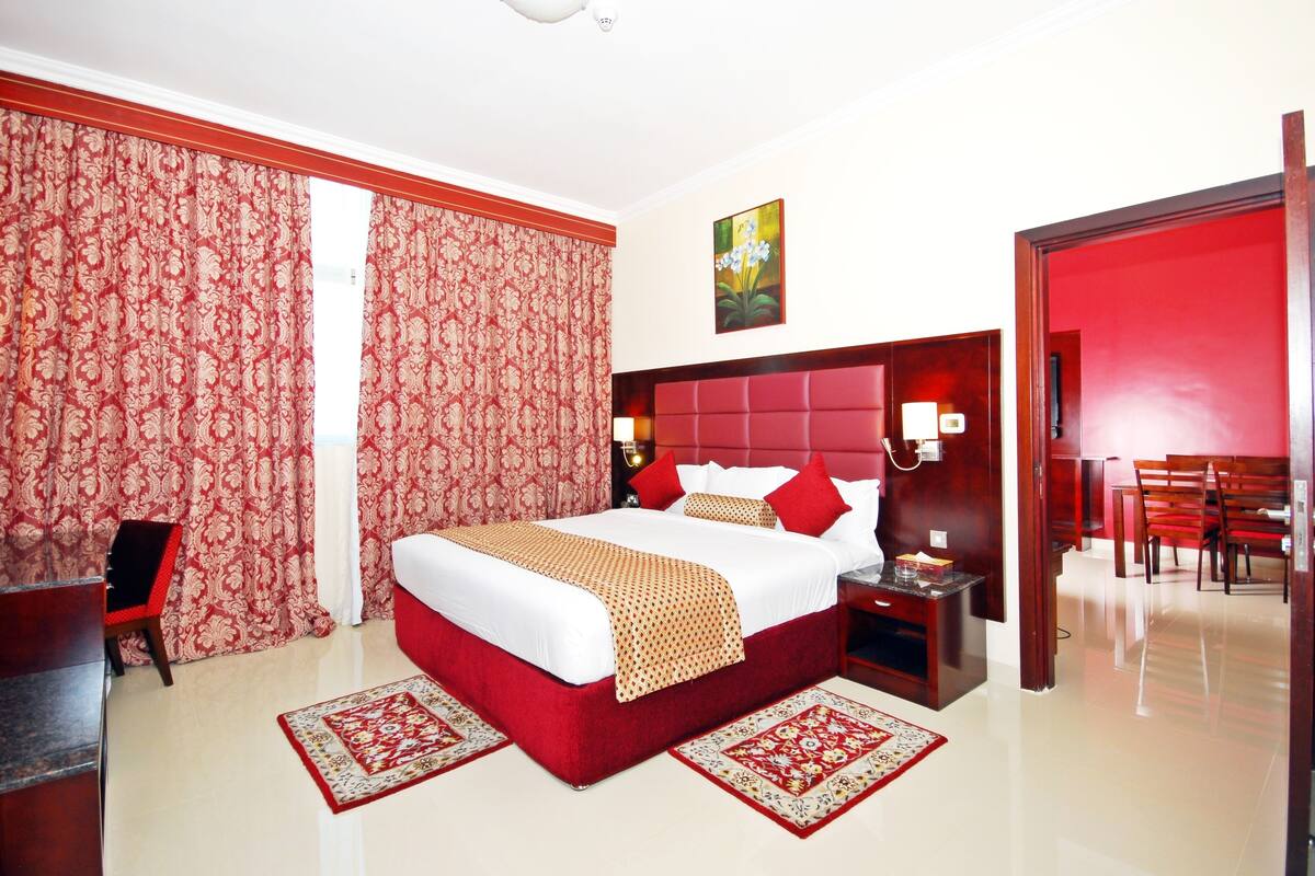 A hotel room with a large bed, red curtains, a wooden headboard, a nightstand, a desk, and a dining area with chairs.