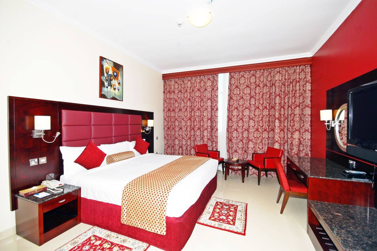 A hotel room with a large bed, red and white curtains, a television, a desk with a chair, and a red carpet.