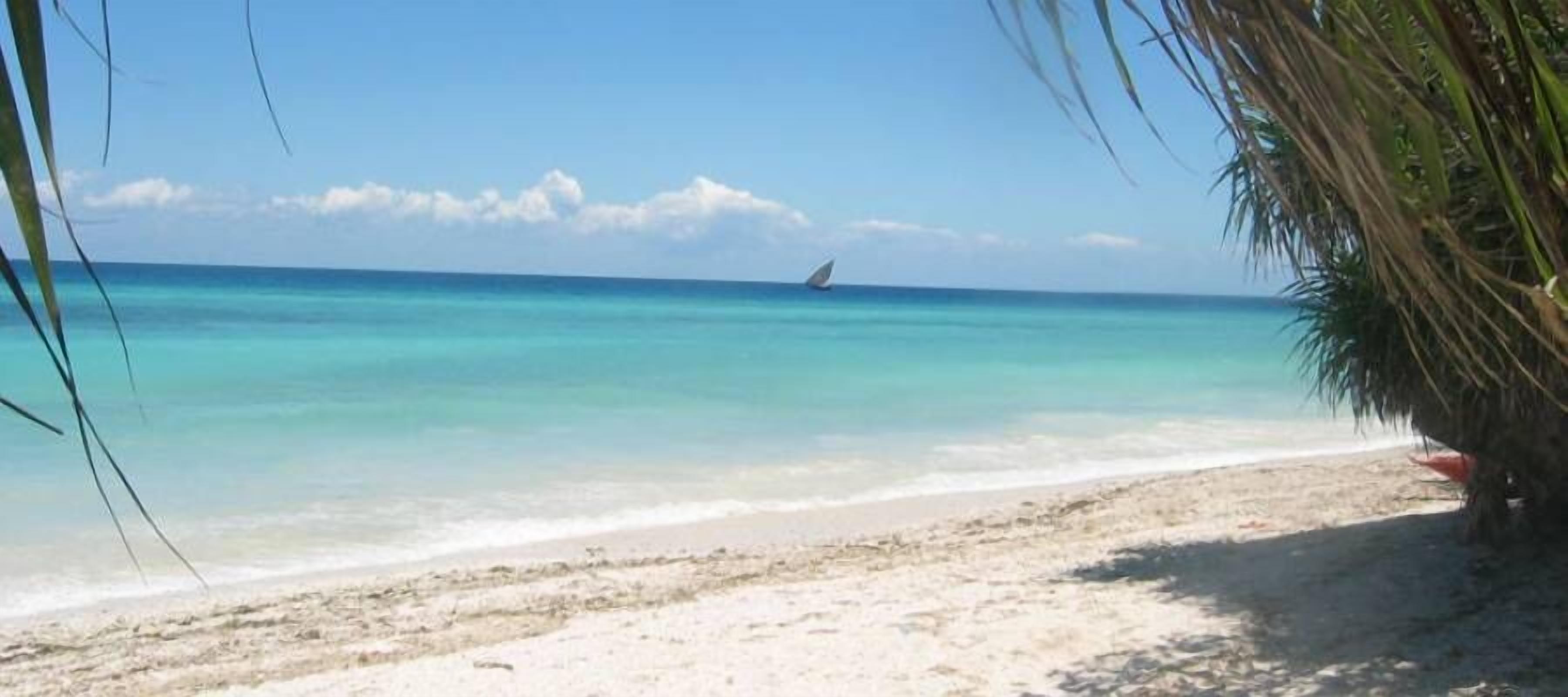 A beach with clear turquoise water and a sailboat in the distance.