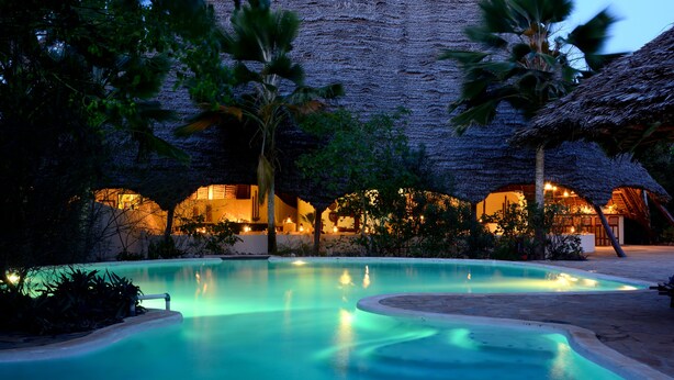 A pool area with a thatched-roof building, palm trees, and a well-lit interior.