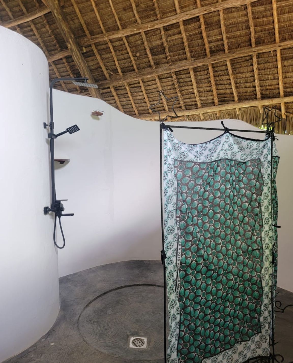 A room with a thatched roof, a shower area with a patterned curtain, and a white wall with a black faucet.