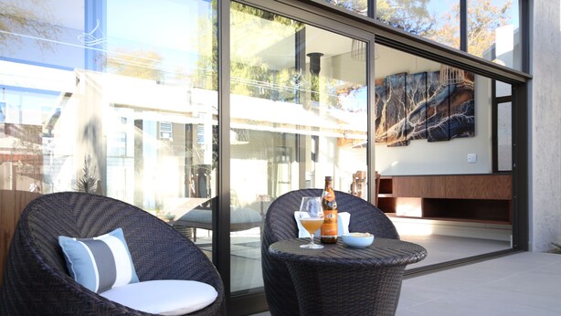 A modern outdoor seating area with wicker chairs and a glass door leading to a patio. - The Elegant Guesthouse (Windhoek)