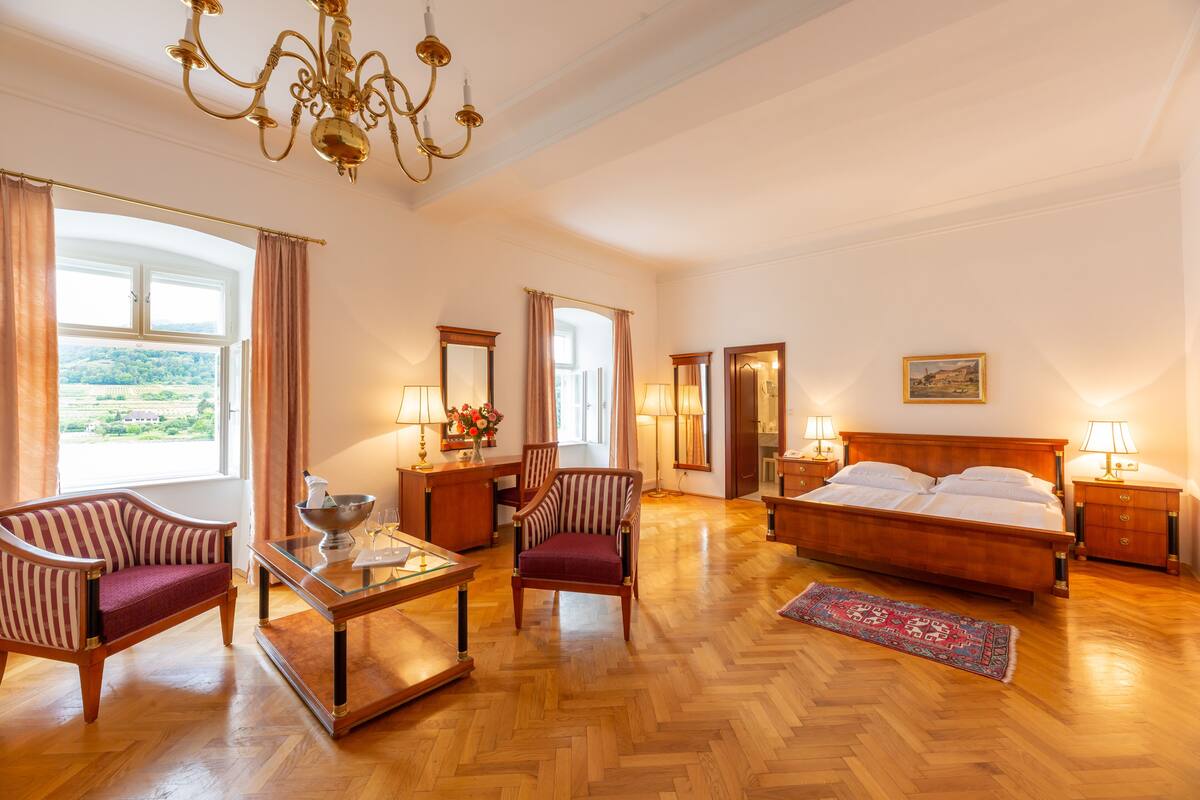 A spacious room with a large bed, a sitting area with a sofa and armchairs, a dining table, and a chandelier.