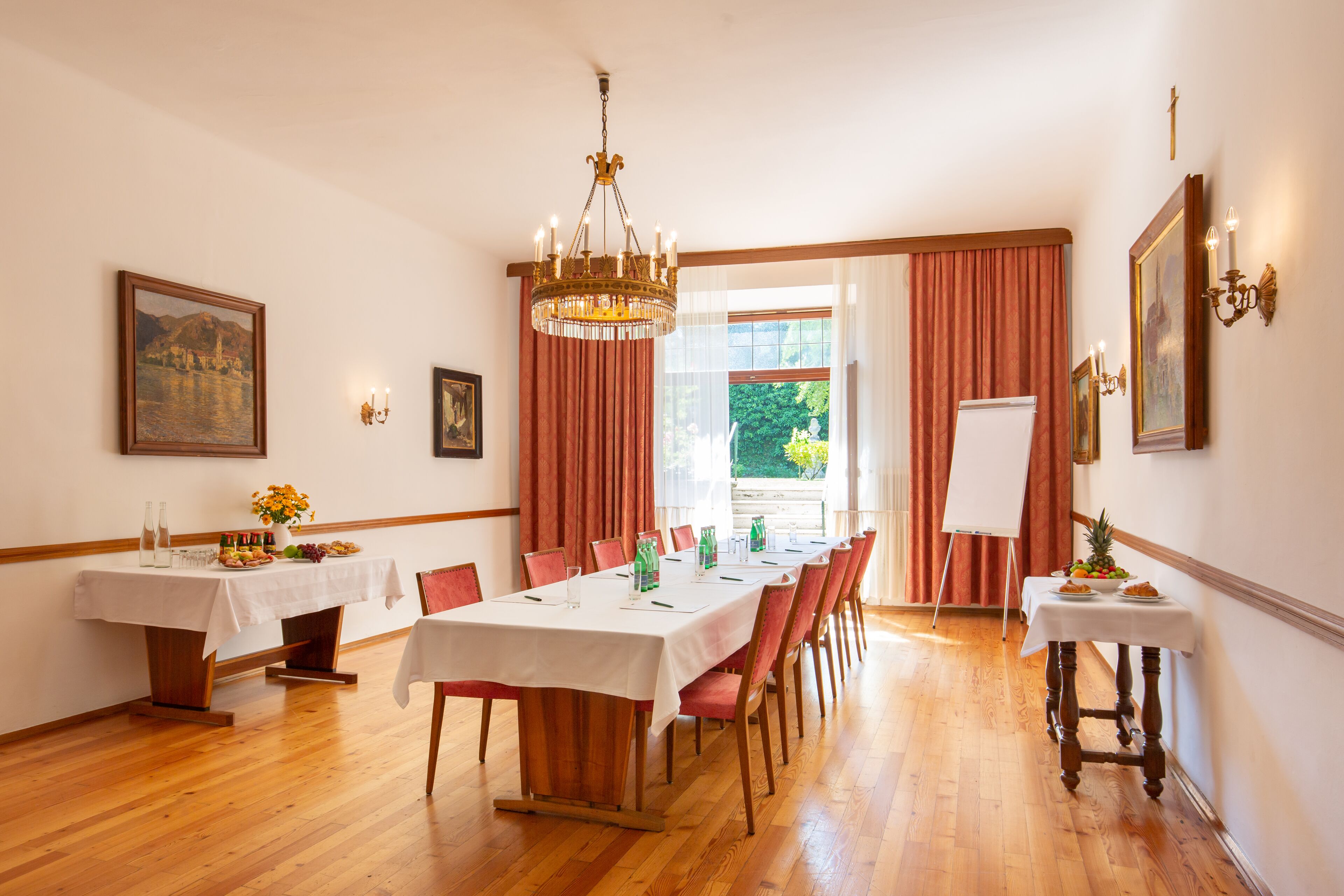 A dining room with a long table set for a meal, surrounded by chairs, with a chandelier above and a painting on the wall.