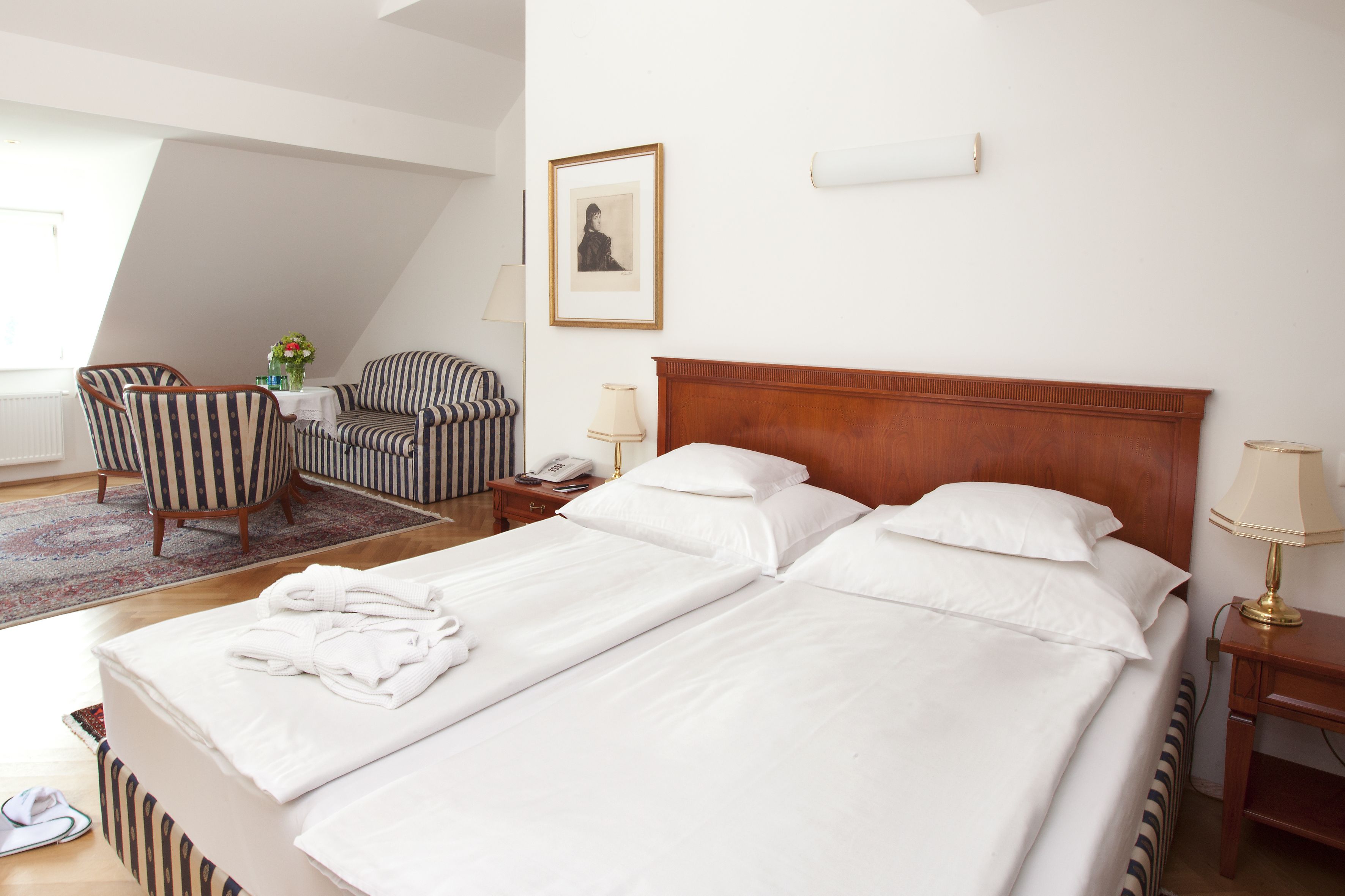 Una habitación de hotel con una cama grande, dos mesitas de noche, una silla, una mesita con un florero y un cuadro en la pared.