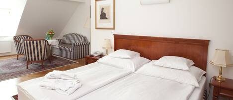 Una habitación de hotel con una cama grande, dos mesitas de noche, una silla, una mesita con un florero y un cuadro en la pared.