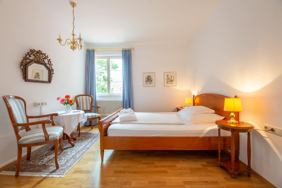 A bedroom with a wooden bed, a nightstand with a lamp, a chair, a small table with flowers, and a window with curtains.