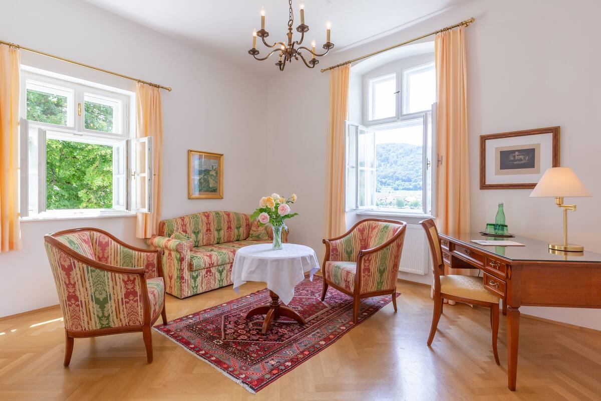 A room with a sofa, chairs, a table, and a desk. There are windows with curtains, a chandelier, and framed pictures on the wall.