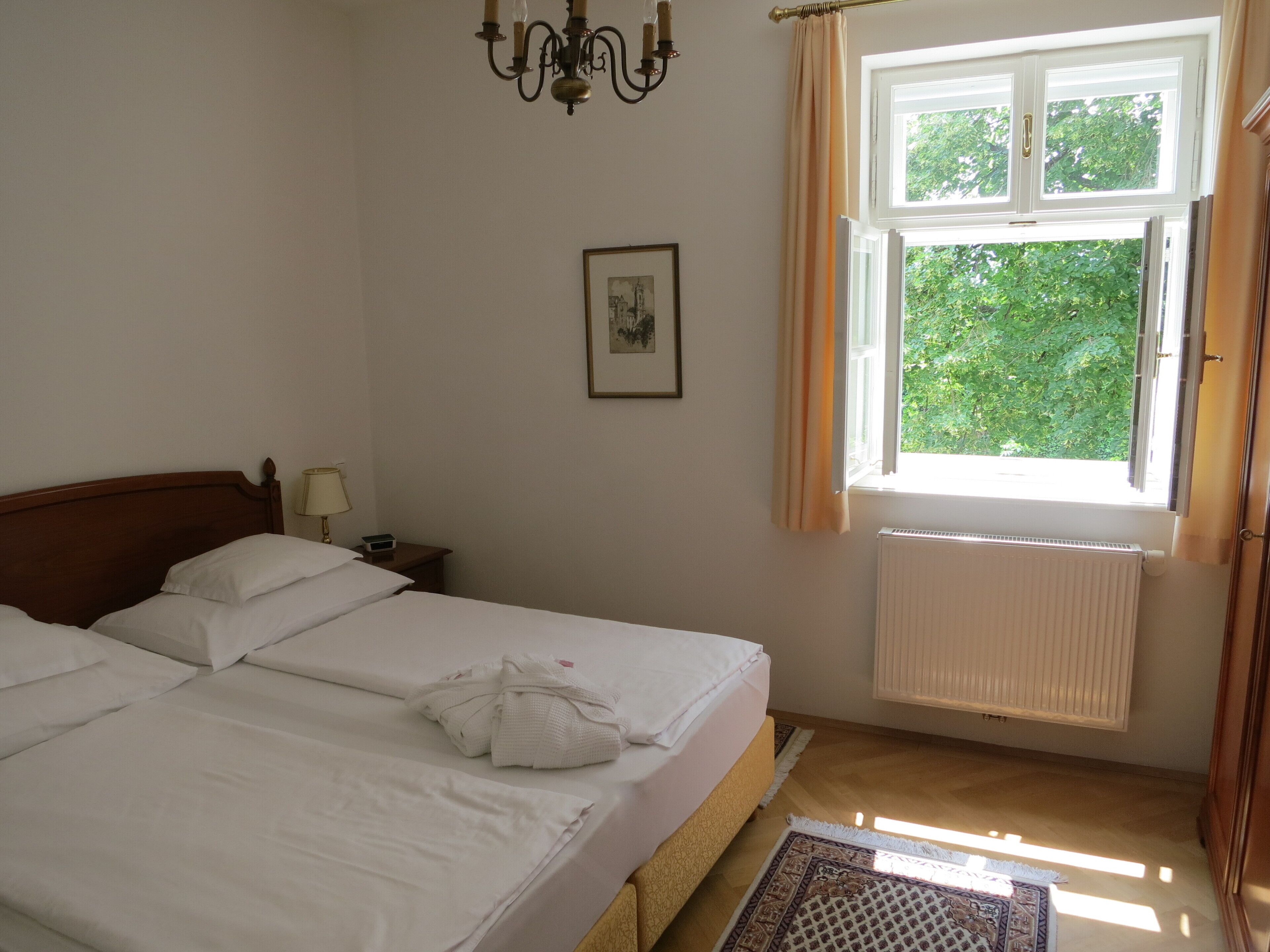 A hotel room with a double bed, a bedside table with a lamp, a framed picture on the wall, a window with curtains, and a radiator.