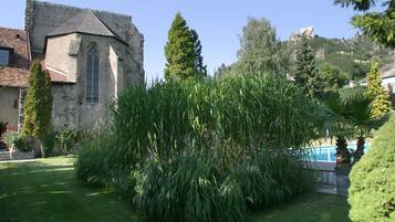 Un bâtiment en pierre doté d’une grande fenêtre et d’un jardin où poussent de hautes herbes et des arbres.