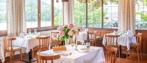 Un comedor con mesas y sillas de madera, centros de mesa con flores y ventanas amplias con cortinas.
