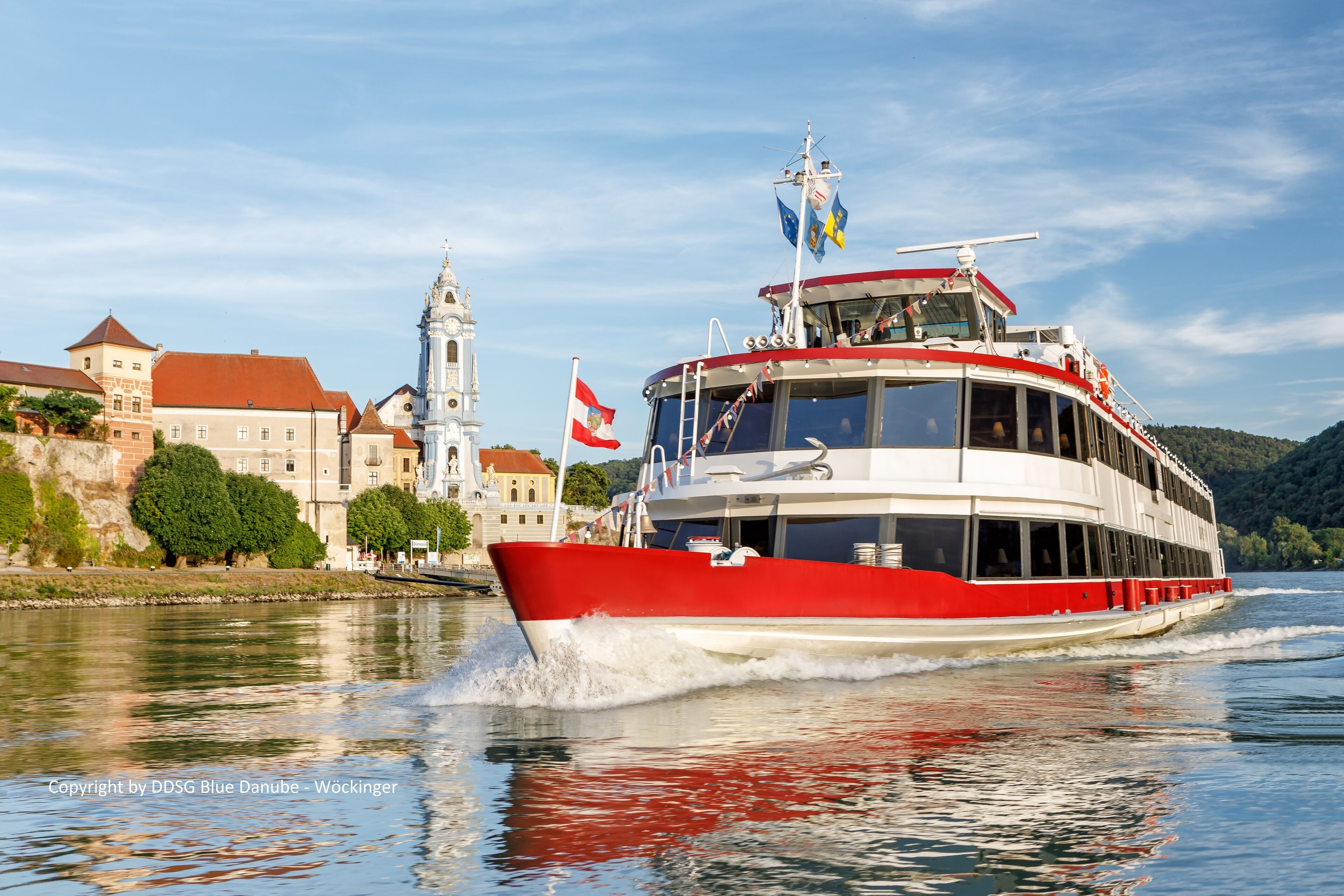 Ein rotes und weißes Boot fährt auf einem Fluss, im Hintergrund ein historisches Gebäude und eine Kirche.