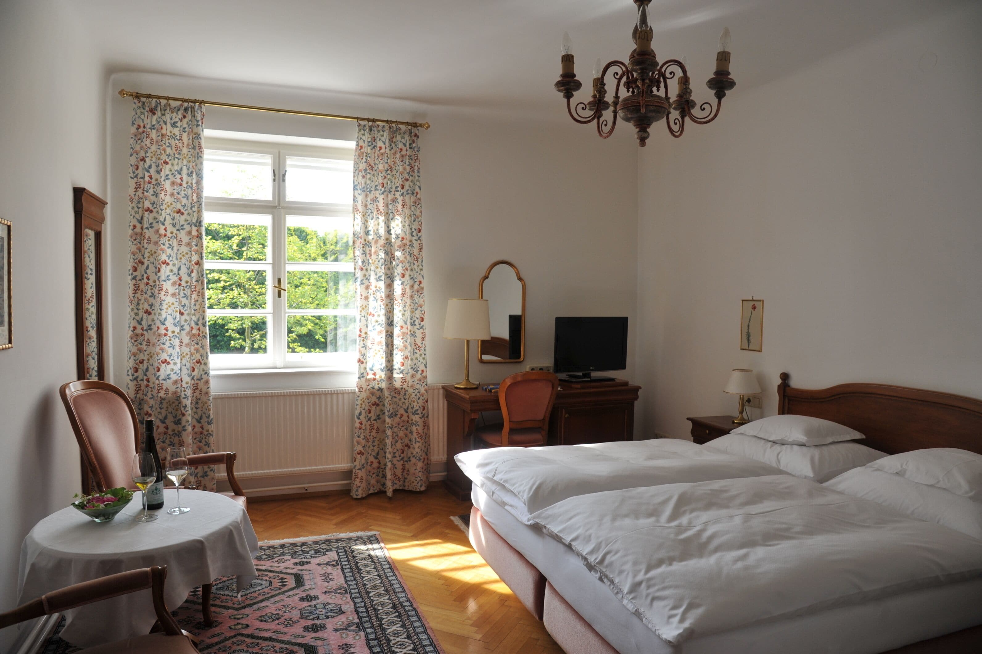 Una camera d'albergo con un letto, una scrivania con un computer, una sedia, un comodino con una lampada e una finestra con tende.