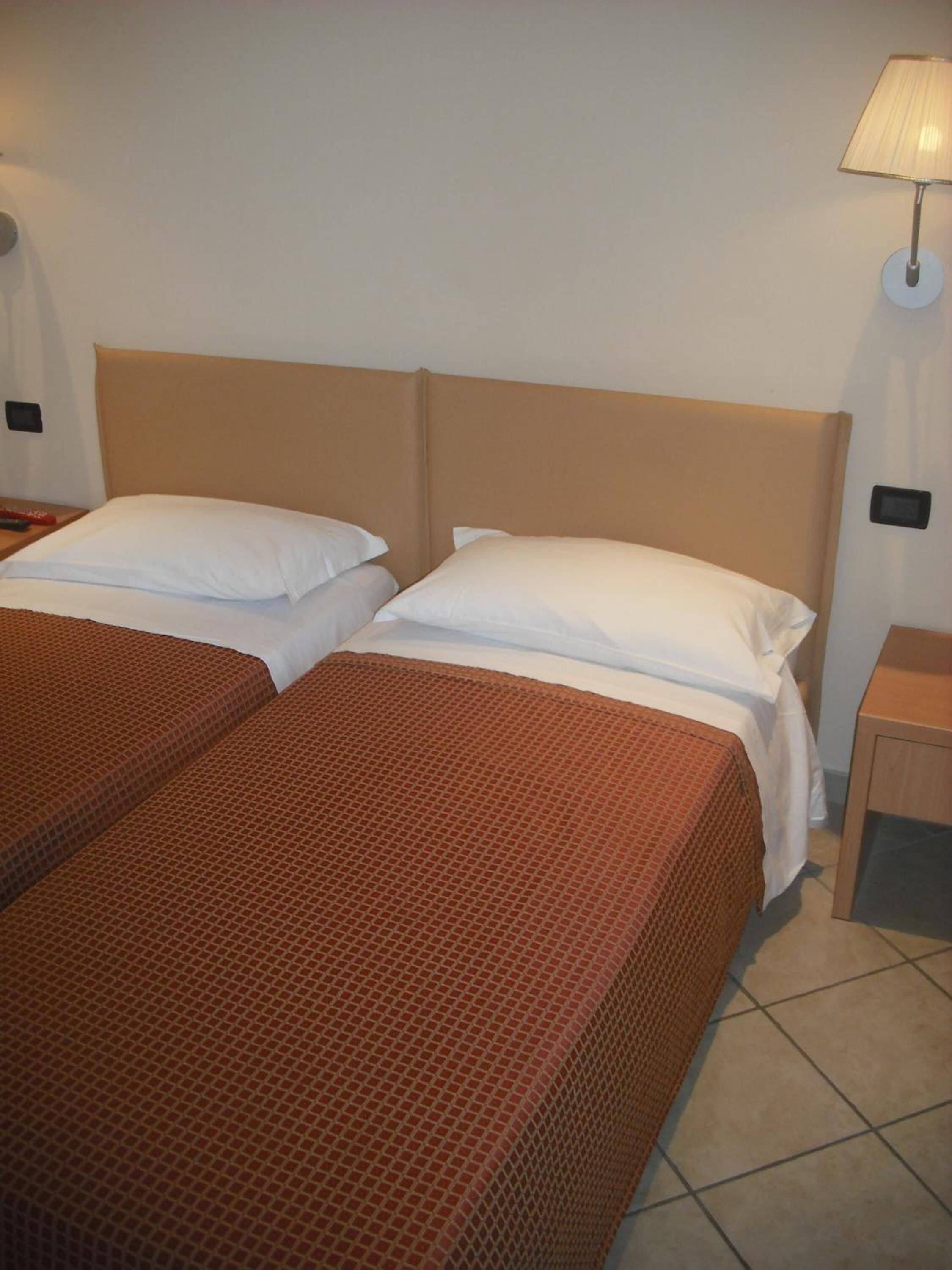 Quarto de hotel com duas camas, cada uma com colcha marrom estampada e travesseiros brancos. Há uma mesa de cabeceira com abajur do lado direito.