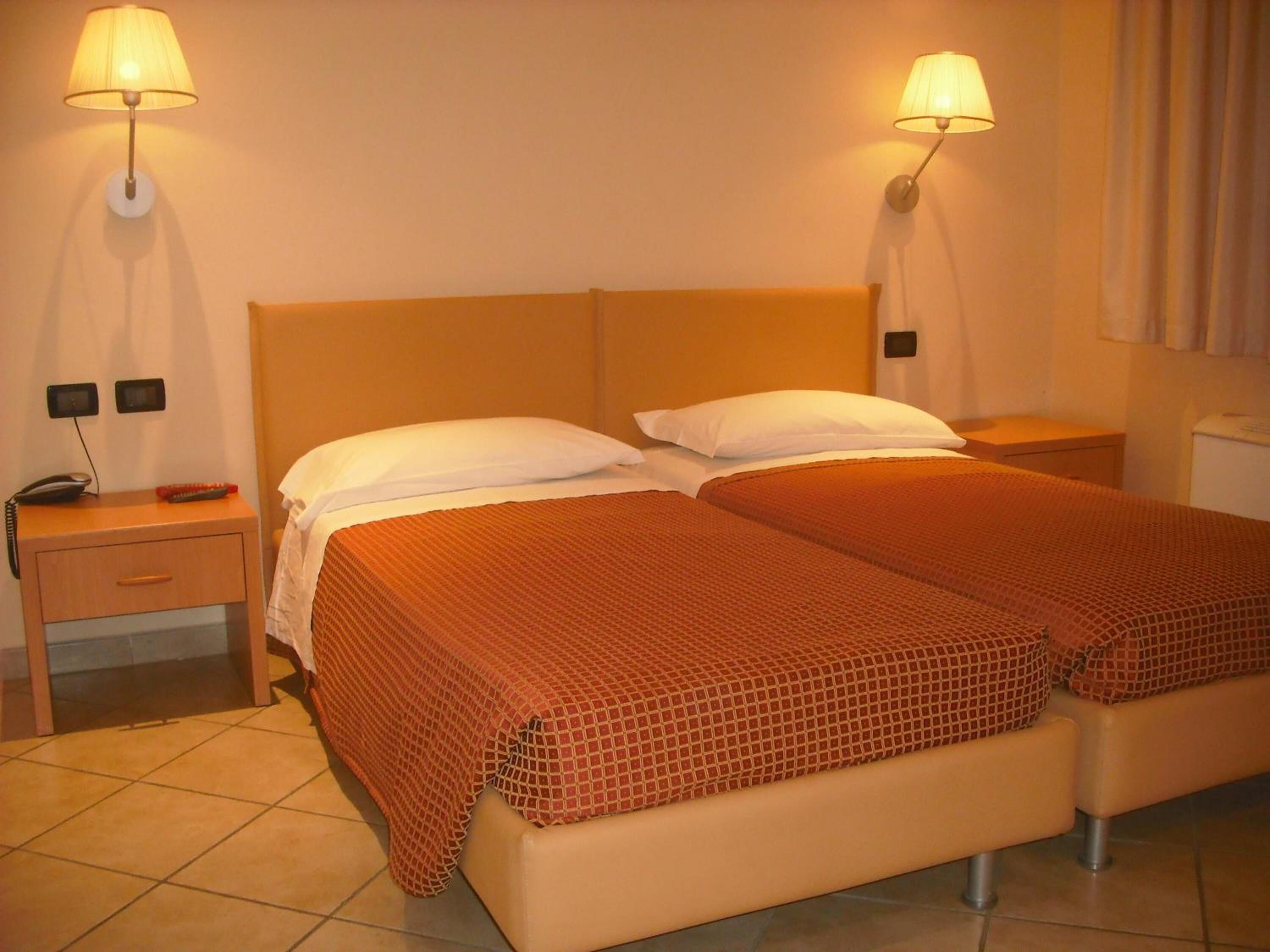 Habitación de hotel con dos camas, cada una con una colcha estampada roja, un cabecero de madera y dos lámparas de pared. Hay una mesita de noche con un teléfono y un objeto rojo encima.
