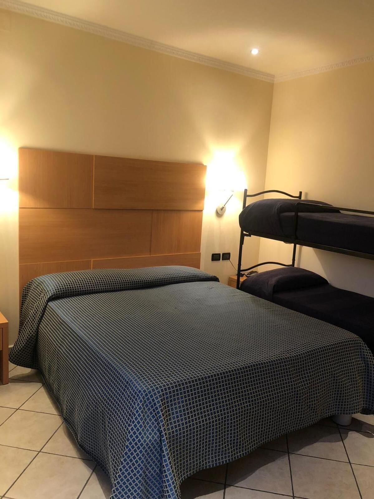 A hotel room with a single bed, a bunk bed, a wooden headboard, and two wall-mounted lights.