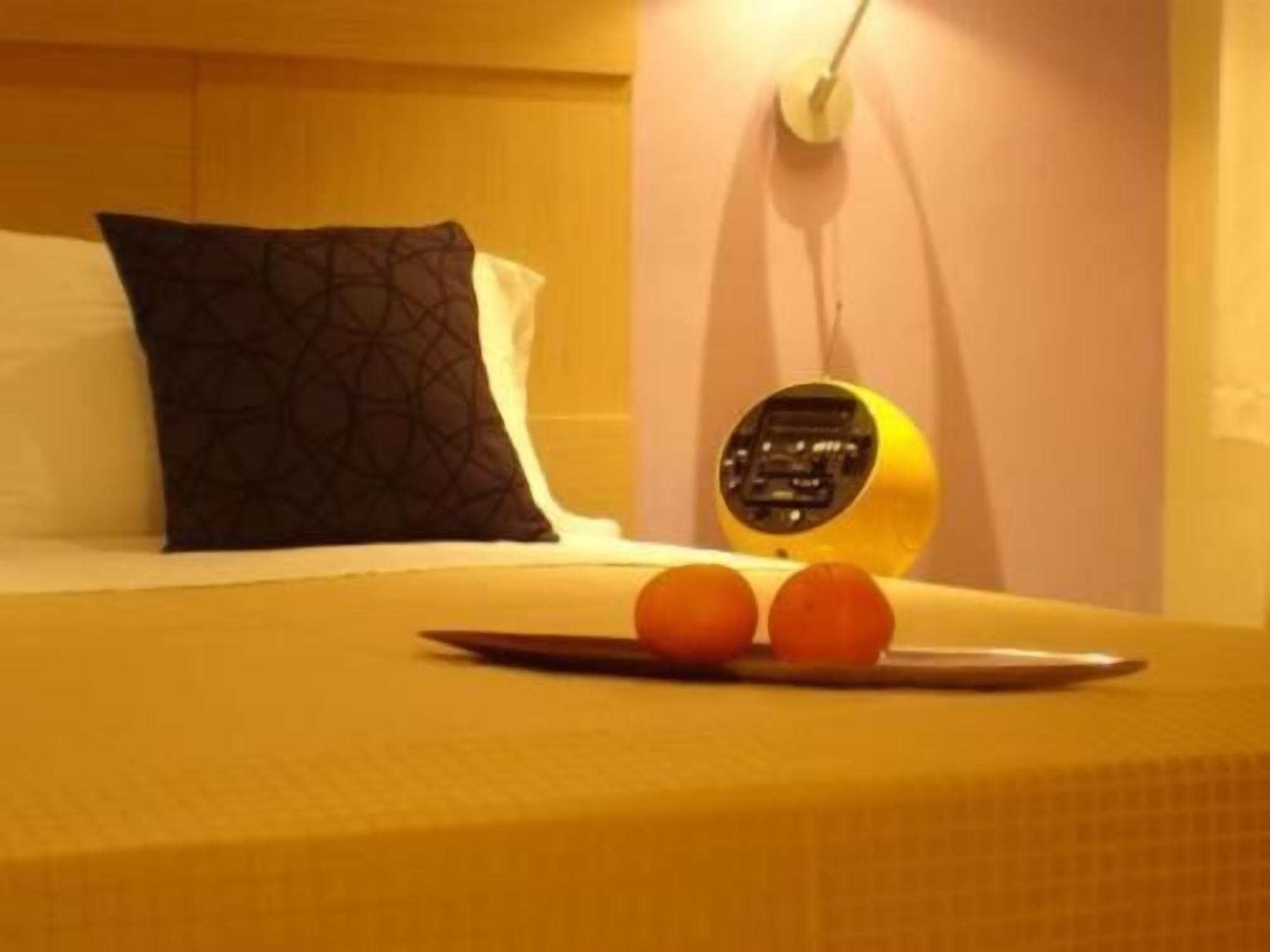 Un lit recouvert d’une couvre-lit jaune, un oreiller noir à motifs et une horloge numérique posée sur une table de chevet.
