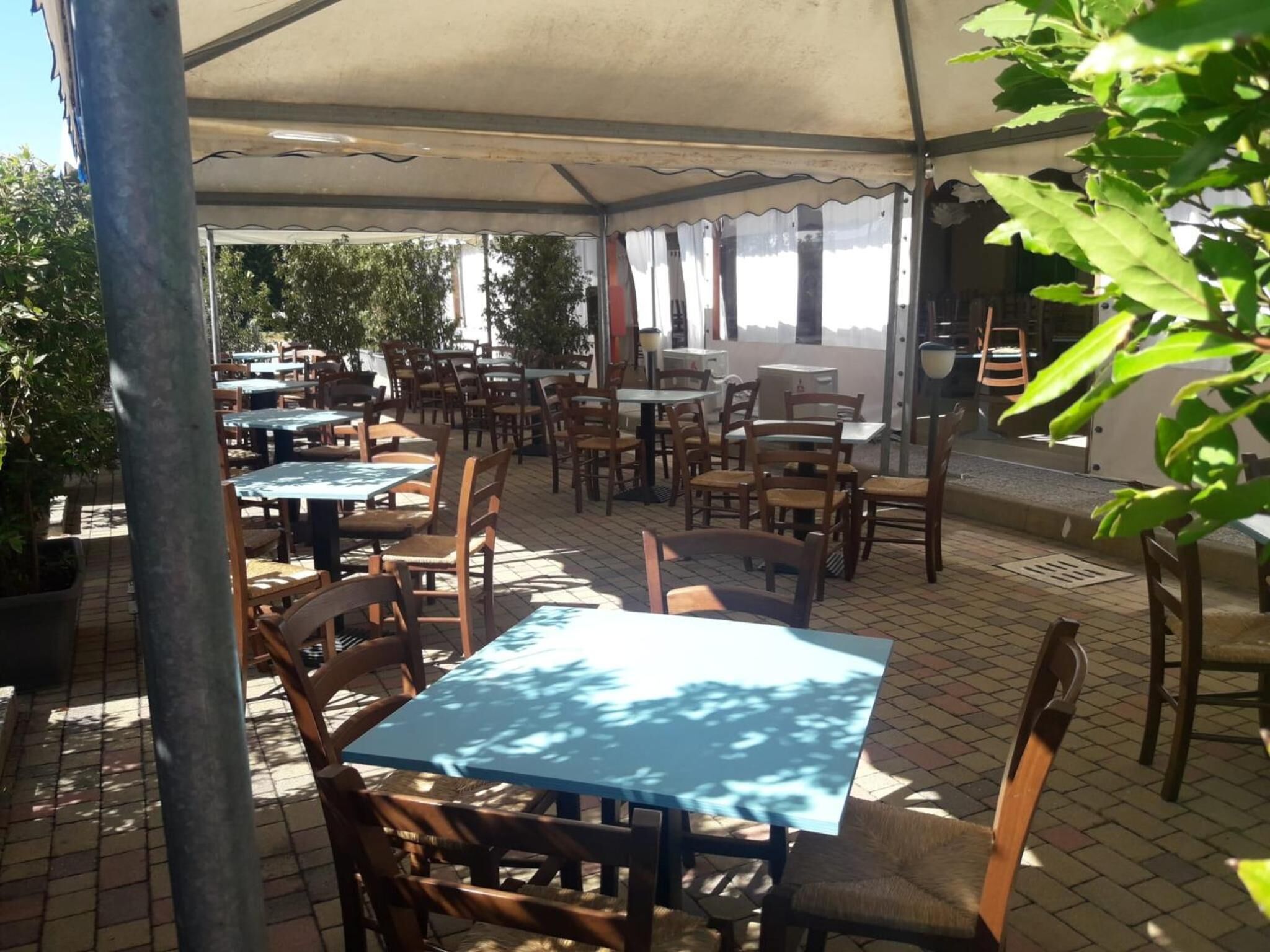 An outdoor cafe with tables and chairs under a canopy, surrounded by greenery.