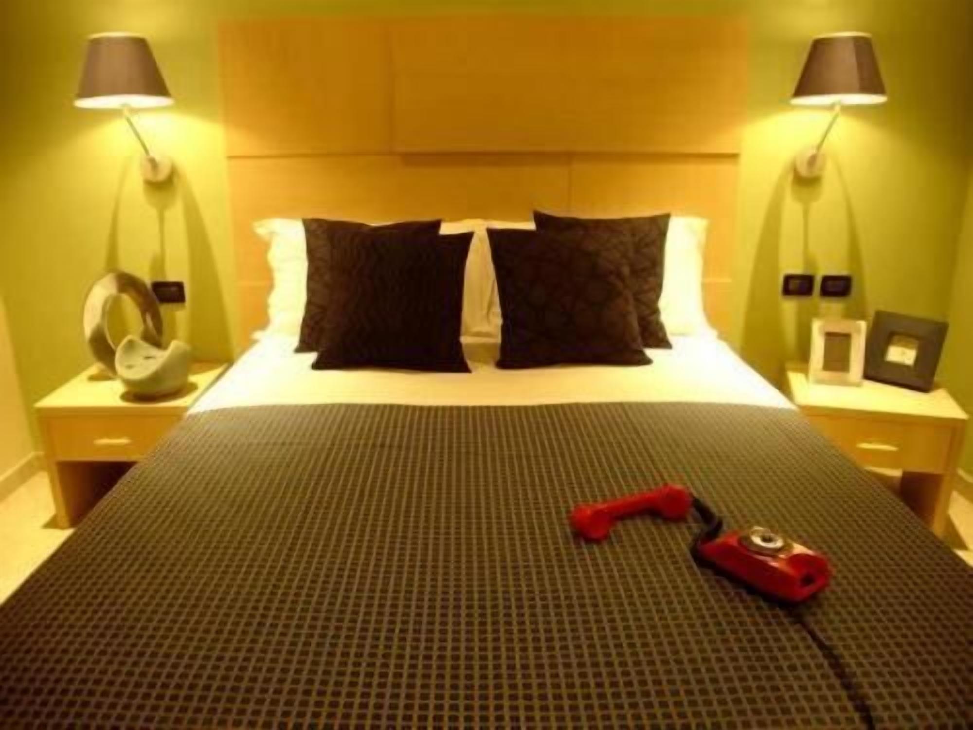 Una habitación de hotel con cama, dos mesitas de noche, una lámpara, un marco de fotos y un objeto rojo sobre la cama.