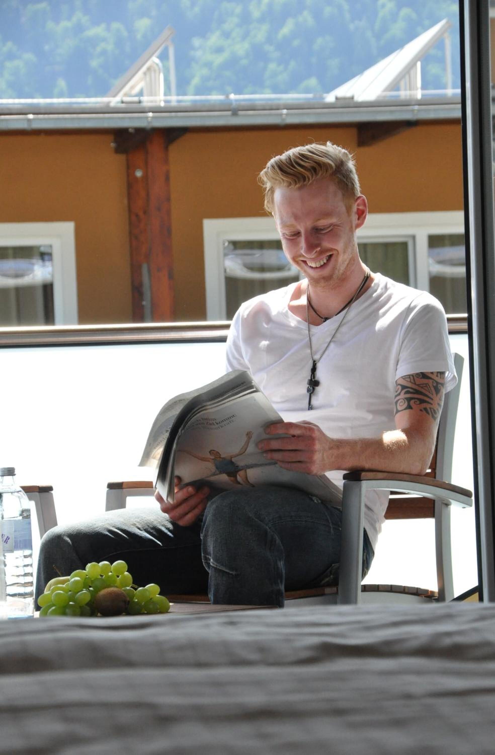 A person sitting on a chair reading a magazine, with a bottle and grapes on the table.