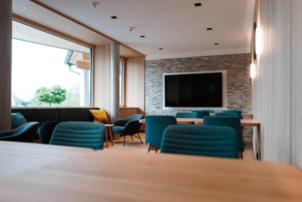 A modern hotel room with a large window, a stone accent wall, a flat-screen TV, and teal upholstered chairs.