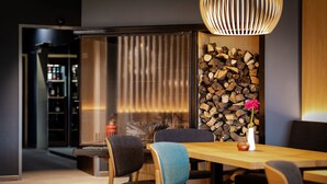 A modern dining area with a wooden table and chairs, a decorative wall with stacked wood, and a hanging light fixture.