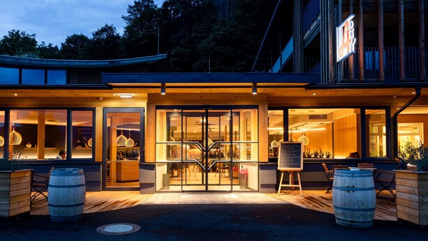 A modern building with large glass doors, wooden barrel seating, and a signboard outside.
