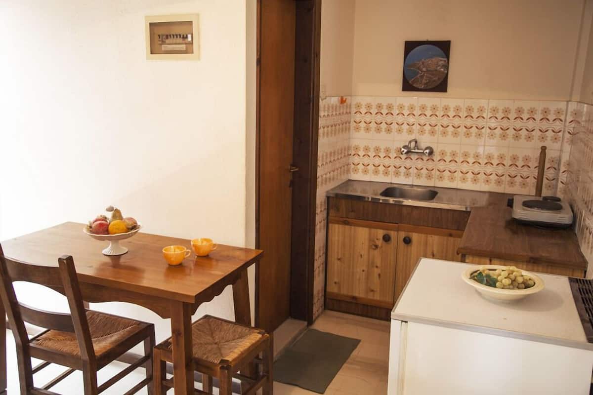 A small kitchen with a wooden table, chairs, a refrigerator, a sink, and a tiled backsplash.