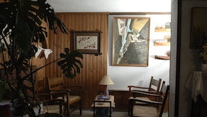 Um quarto acolhedor com painéis de madeira, uma janela grande e uma pintura de uma cena de rua. Há três cadeiras de madeira e uma mesinha com um abajur.